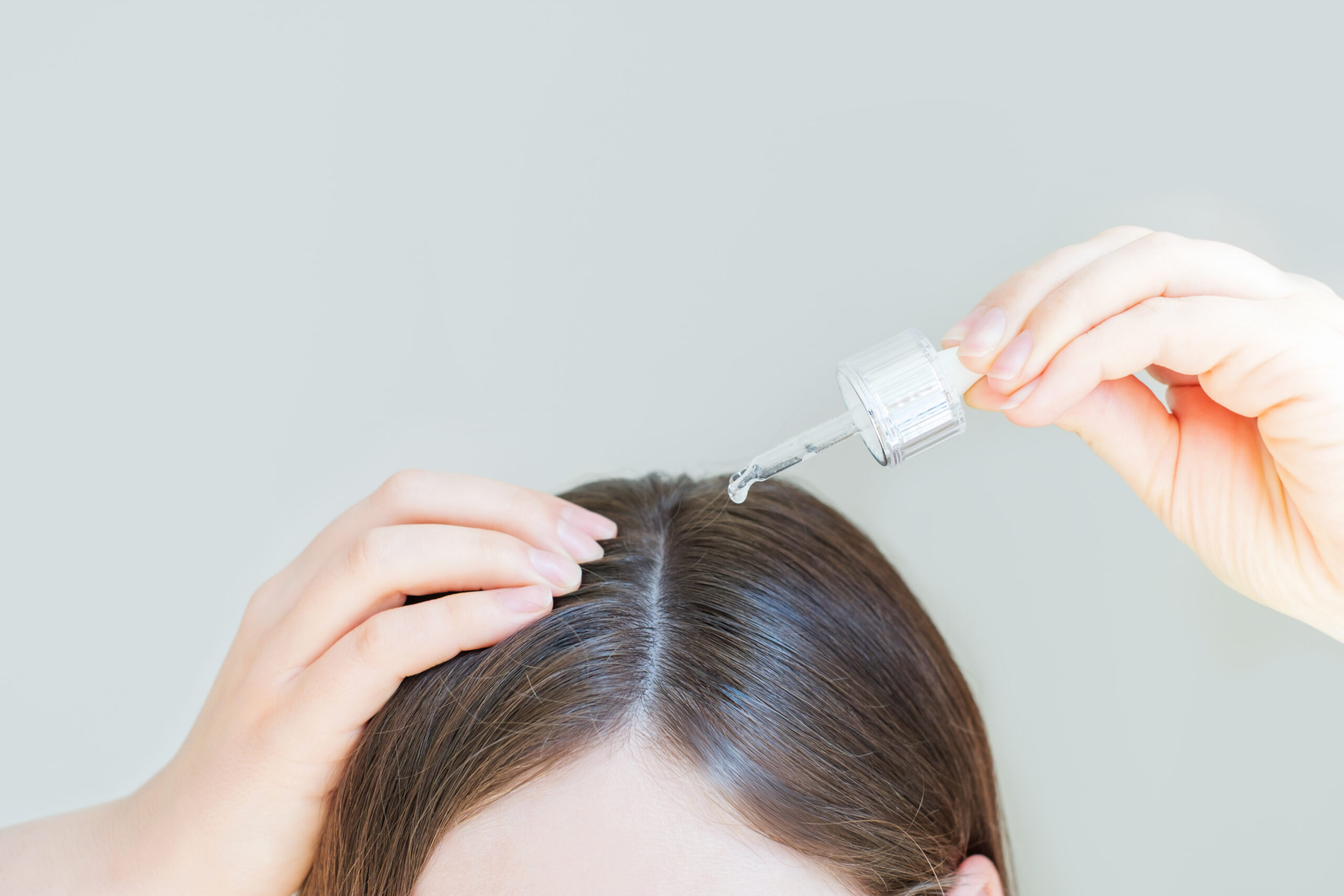 Young woman applying oil to her hair with a pipette on a gray background.